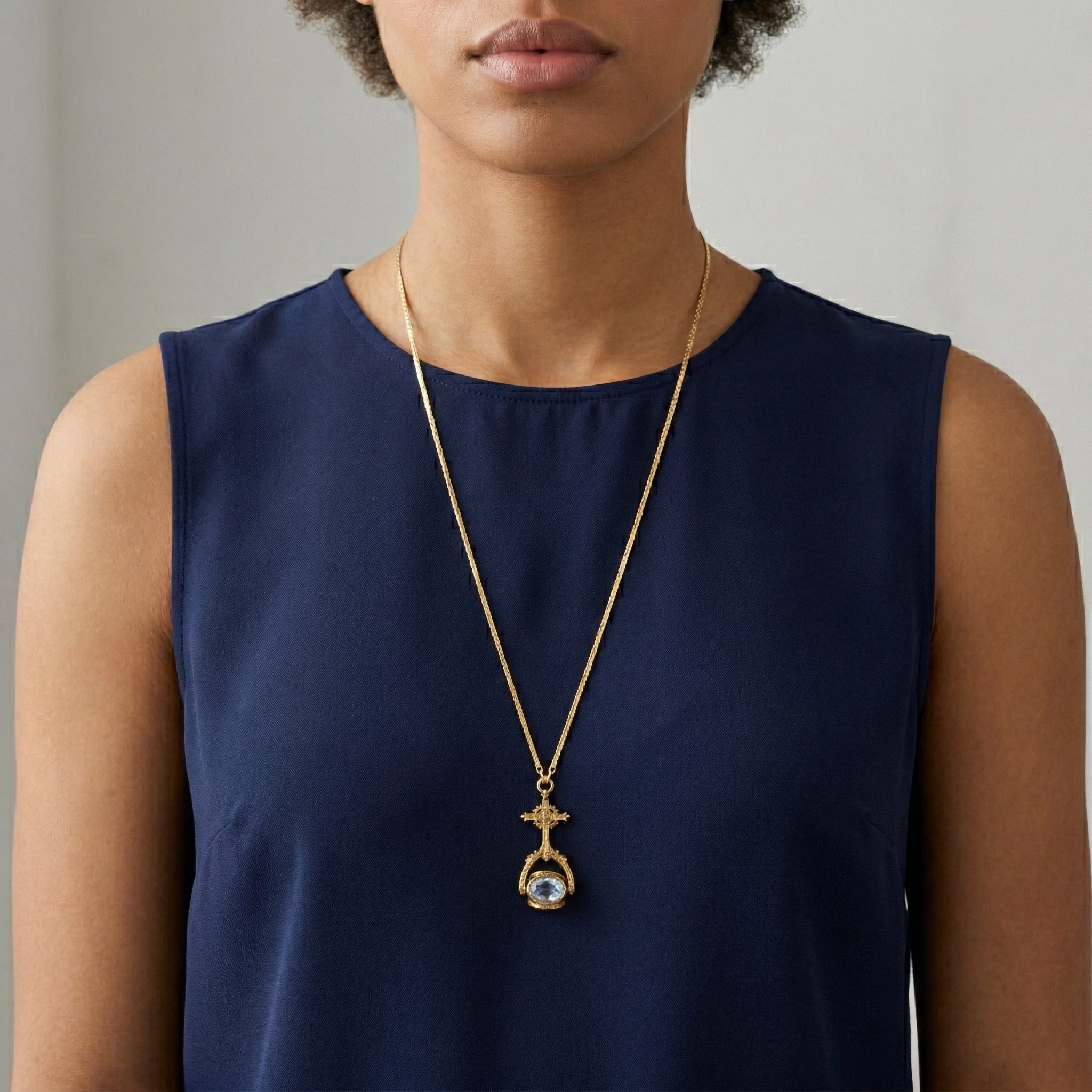Woman wearing a gold necklace with a blue crystal spinner pendant on a plain background