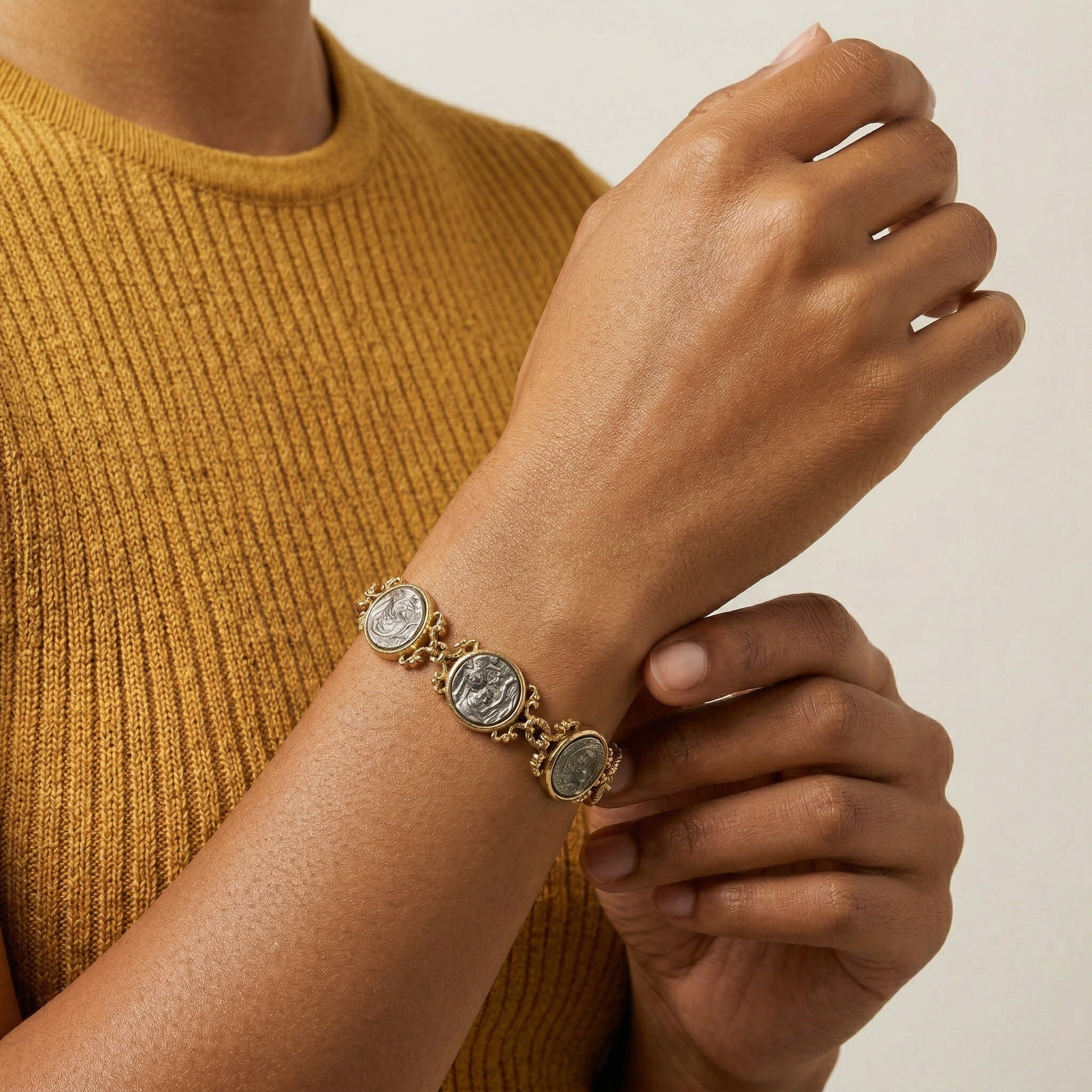 Person wearing a gold bracelet with circular silver tone Mother Mary and Child charms on a beige background