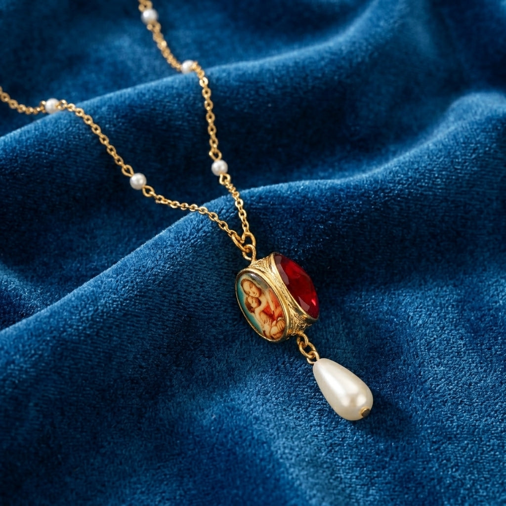Gold necklace that has faux pearl accents with a red crystal pendant and pearl drop on a blue fabric background