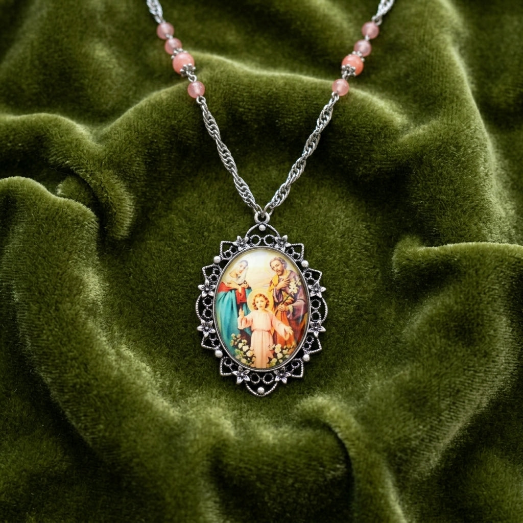 Holy Family pendant necklace with raspberry beads resting on a dark green fabric surface. 