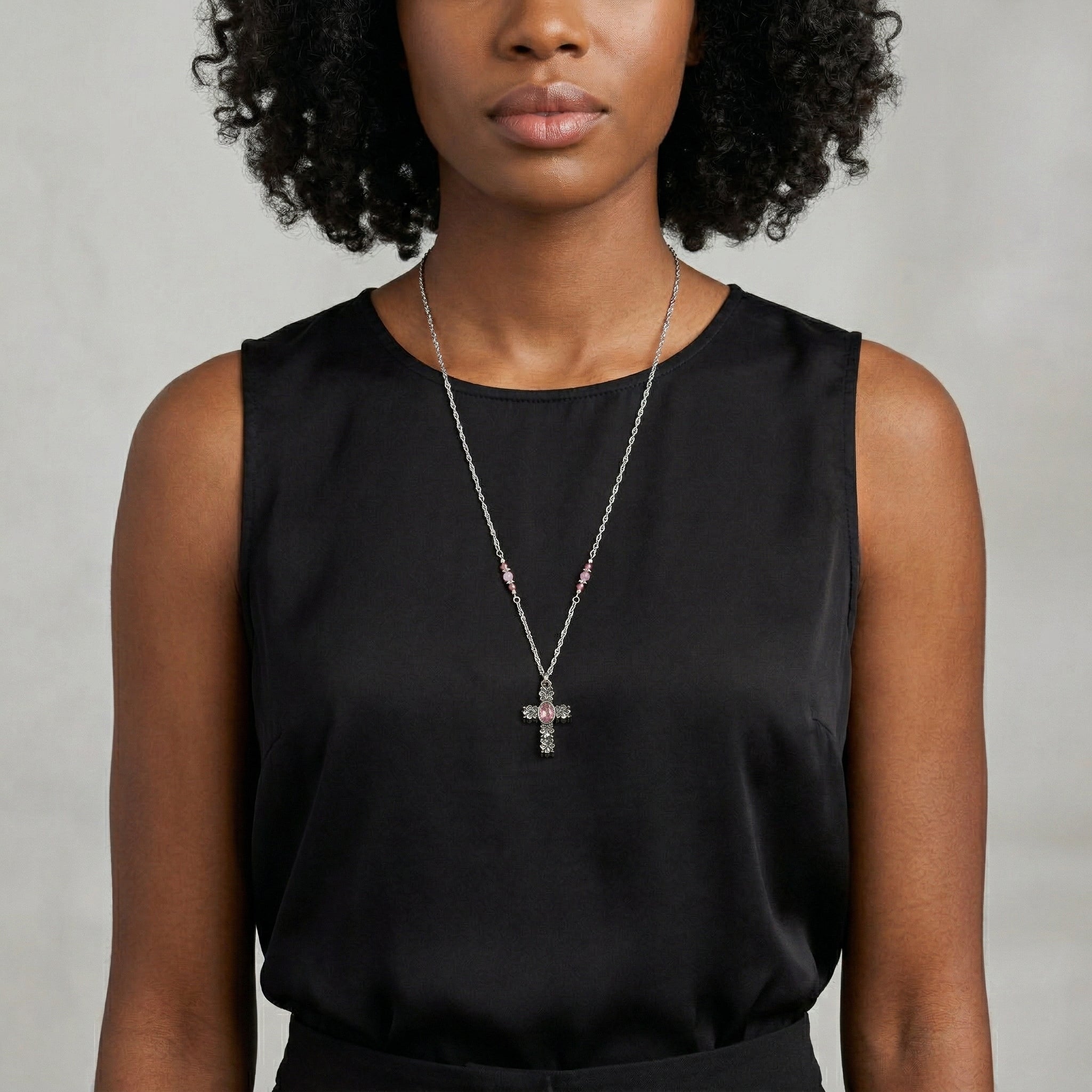Woman wearing a black sleeveless top and a necklace with a cross pendant and a pink crystal against a neutral background