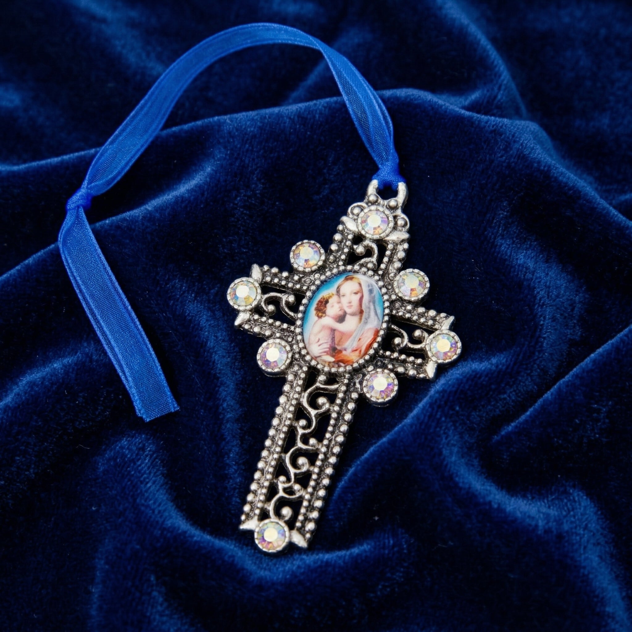 Decorative cross ornament with a blue ribbon and Mother Mary and Child design on a blue velvet background