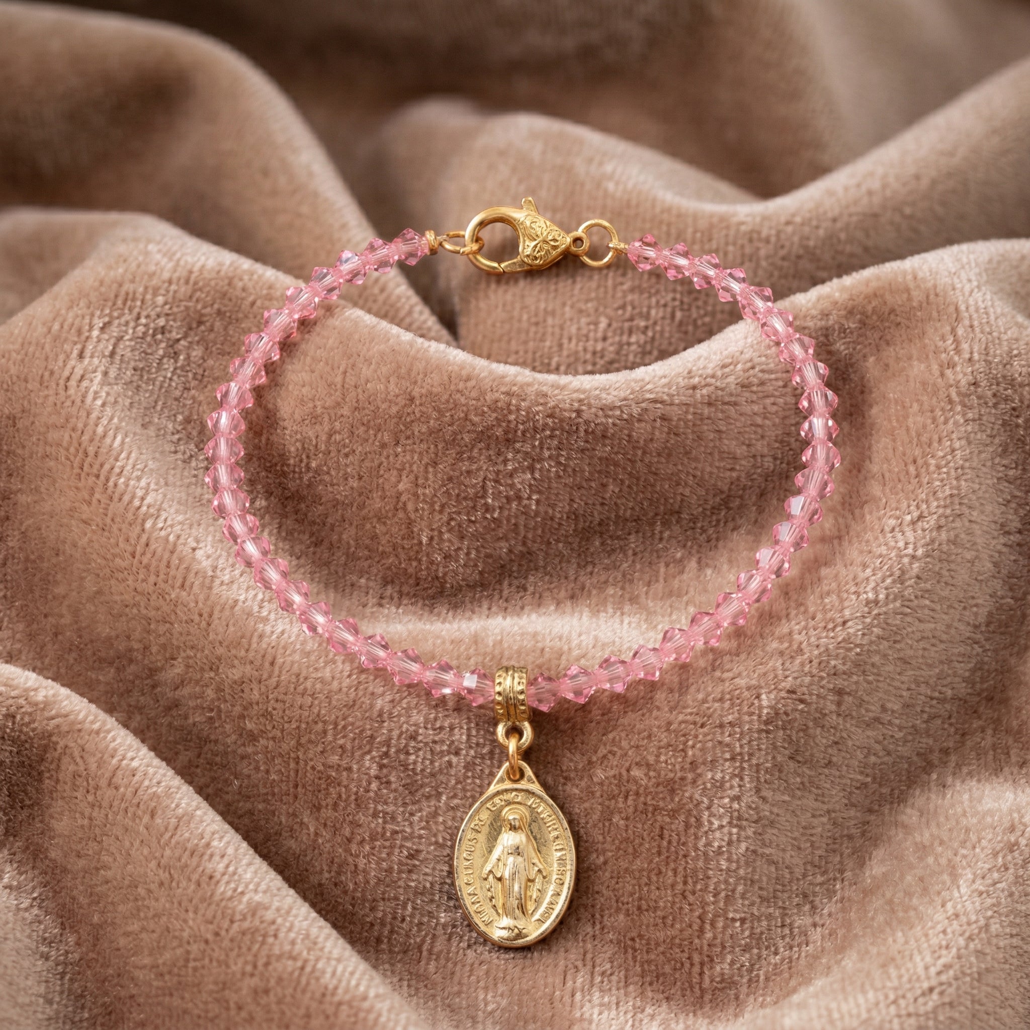 Pink beaded bracelet with gold Jesus charm and pendant on a brown fabric background