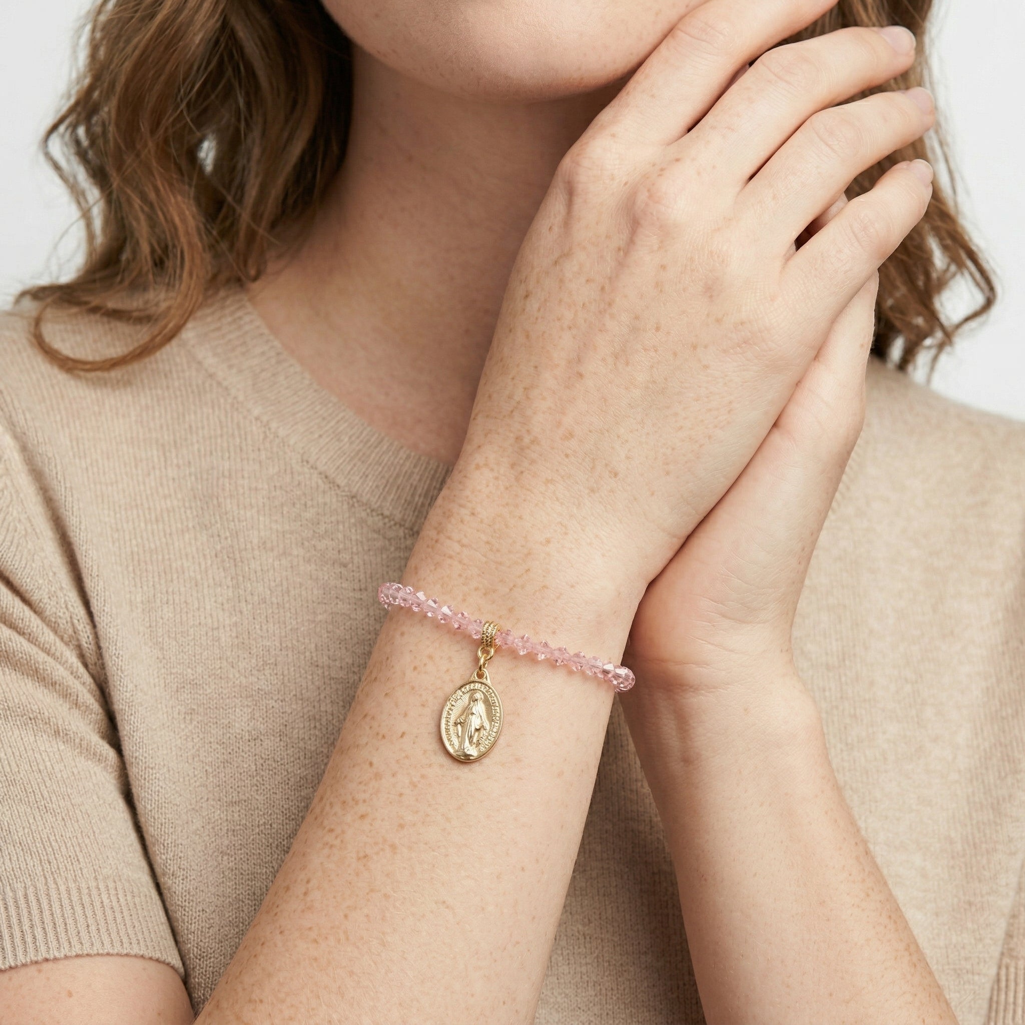 Person wearing a pink beaded bracelet with a gold Jesus charm on a neutral background