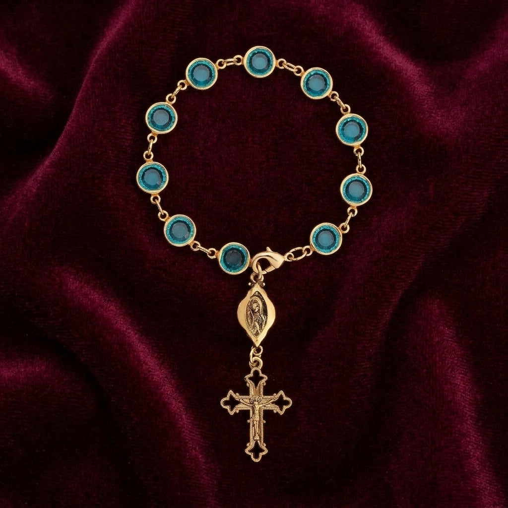 Gold rosary cross charm bracelet with blue crystals on a maroon fabric background