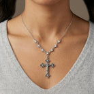 Silver necklace that has crystals on the chain with a cross pendant worn by a person in a gray sweater.