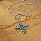 Silver cross pendant with green stone on a chain against a brown stone background