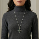 Person wearing a long silver tone cross necklace with a green stone pendant on a plain background