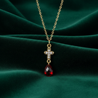 Gold necklace with a red teardrop and cross pendant on a green fabric background