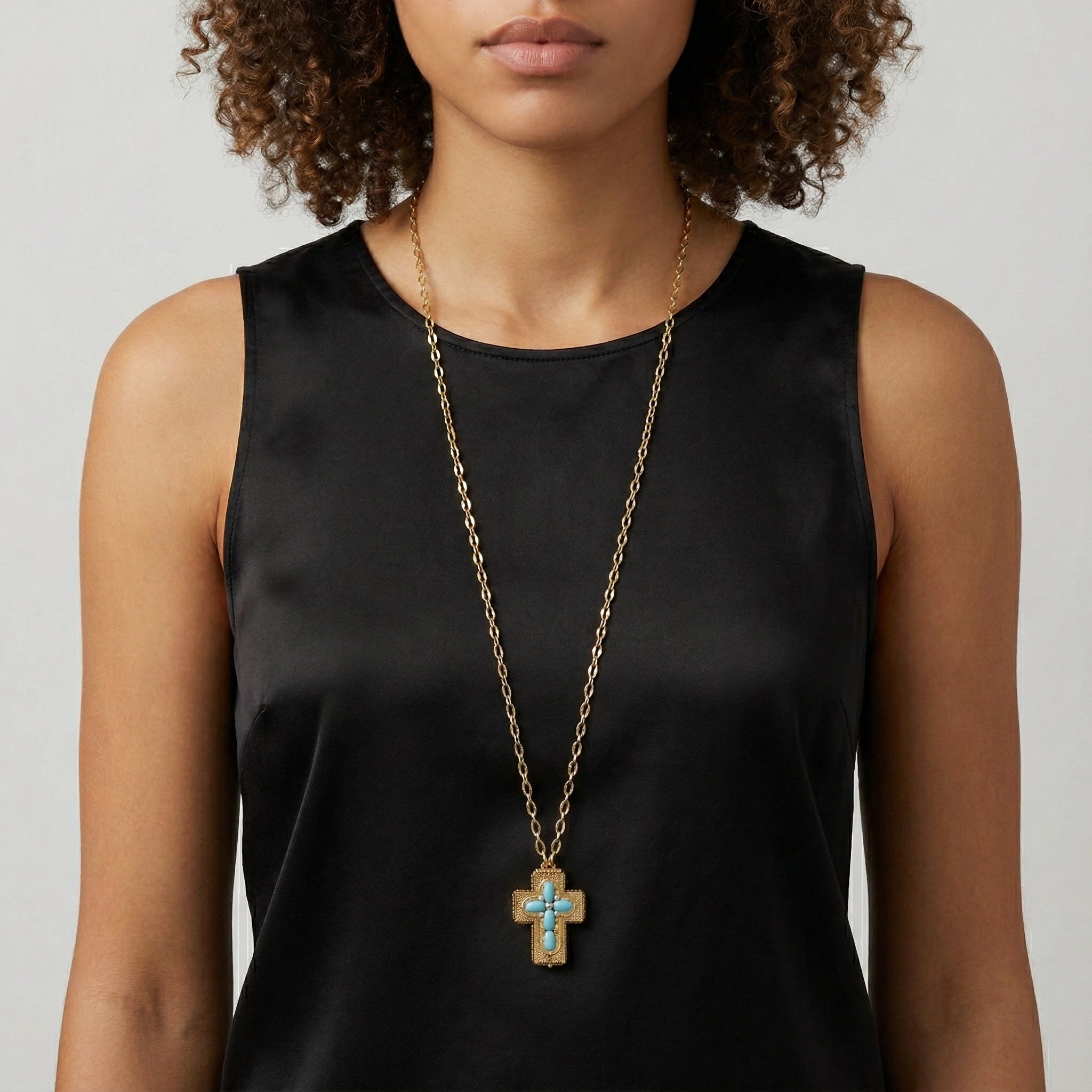 Woman wearing a black sleeveless top with a gold necklace featuring a cross pendant with blue stones. 