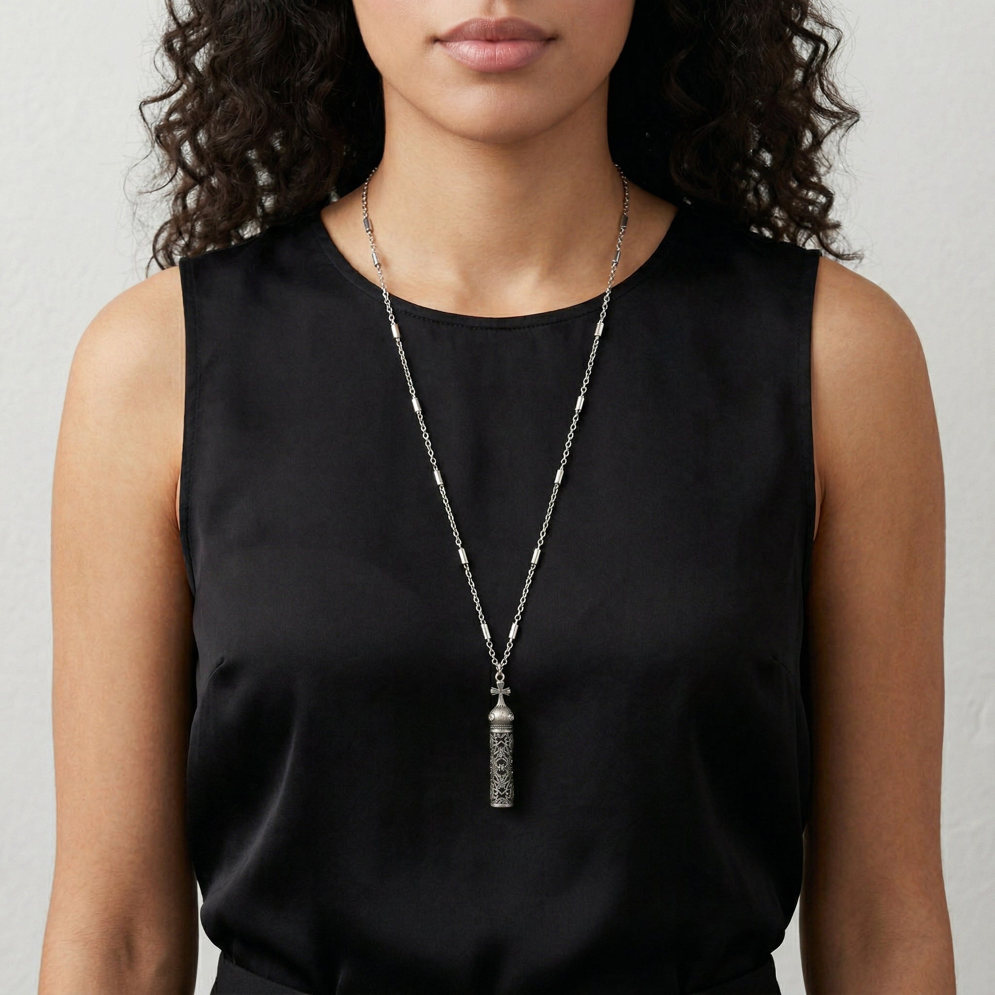 Woman wearing a black sleeveless top with a long vial pendant necklace with a cross design. 
