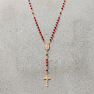 Gold tone rosary cross with red beads resting neatly on a cement surface. 