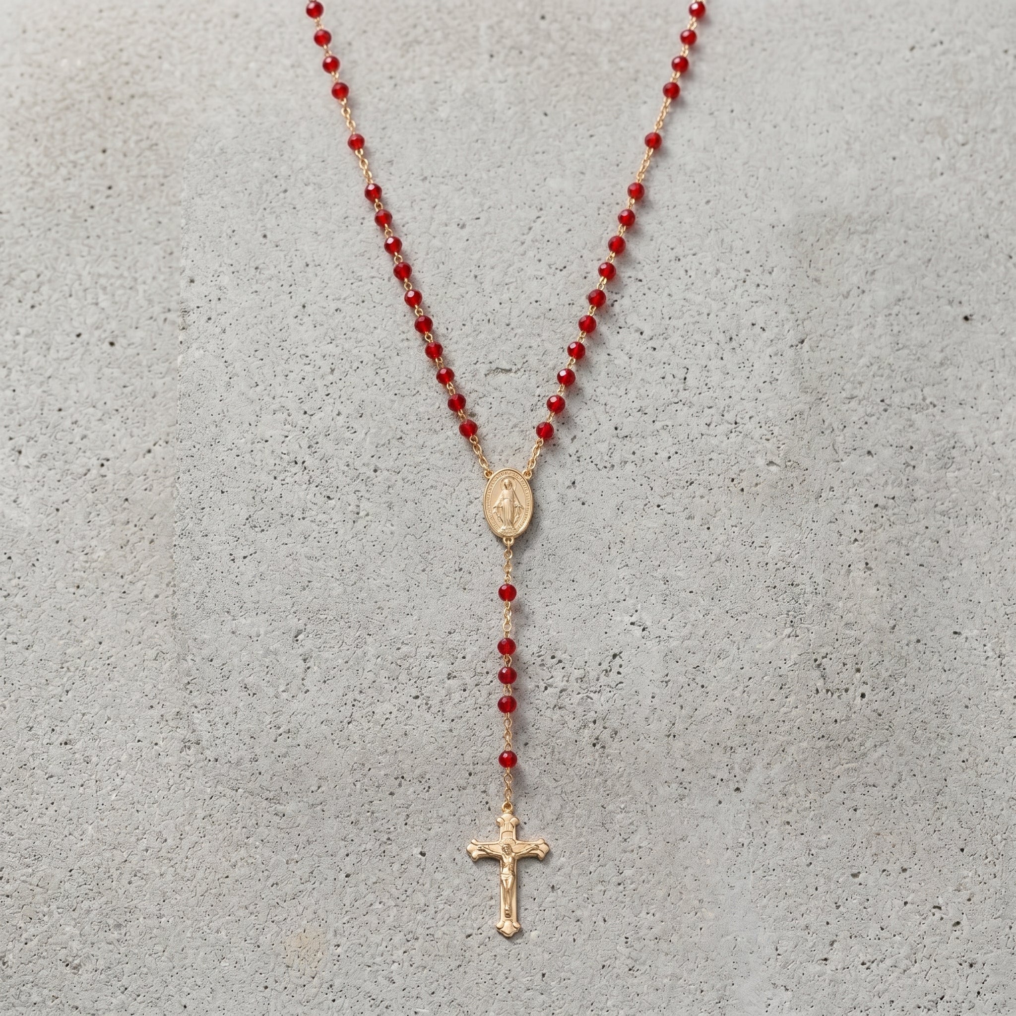 Gold tone rosary cross with red beads resting neatly on a cement surface. 