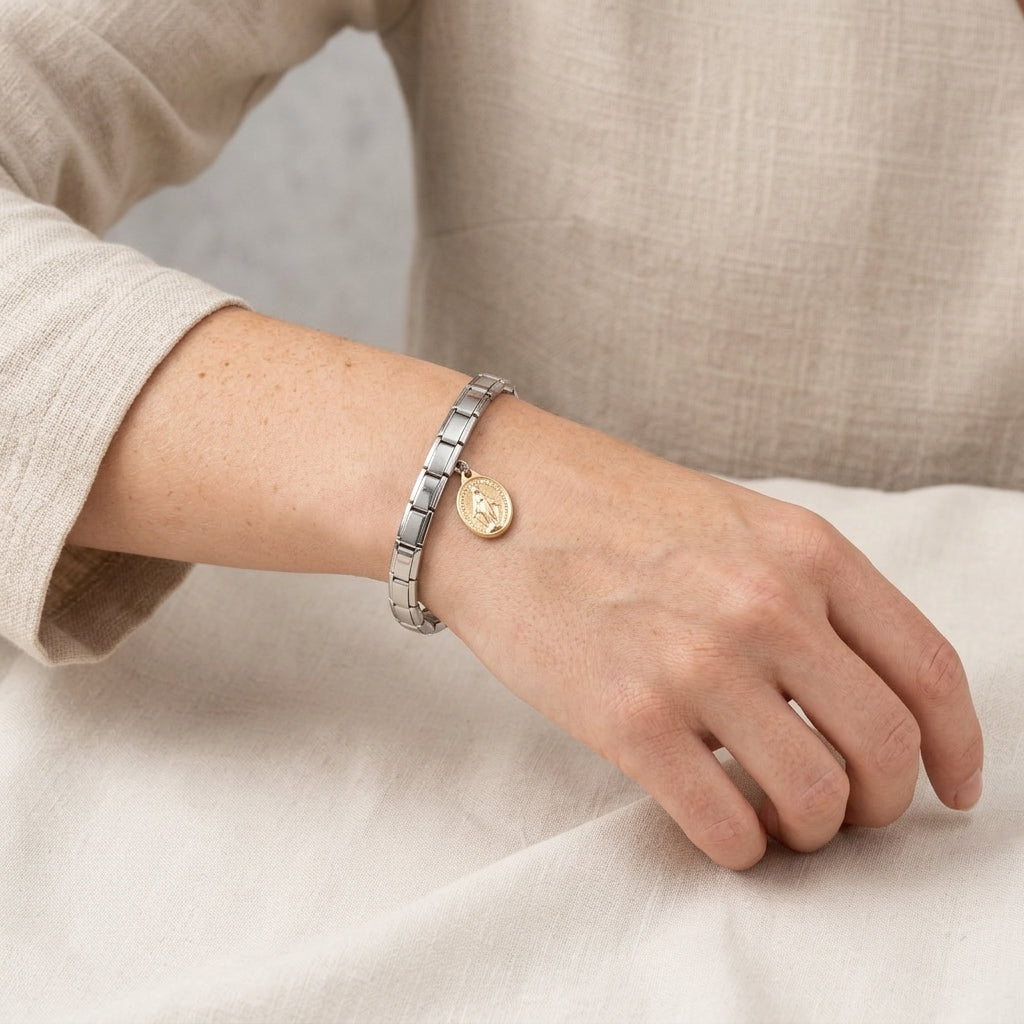 Close-up of a wrist wearing a silver bracelet with a gold Mother Mary charm on a neutral background