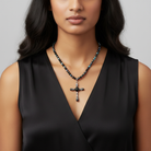 Woman wearing a black dress with a necklace featuring a cross pendant on a gray background.