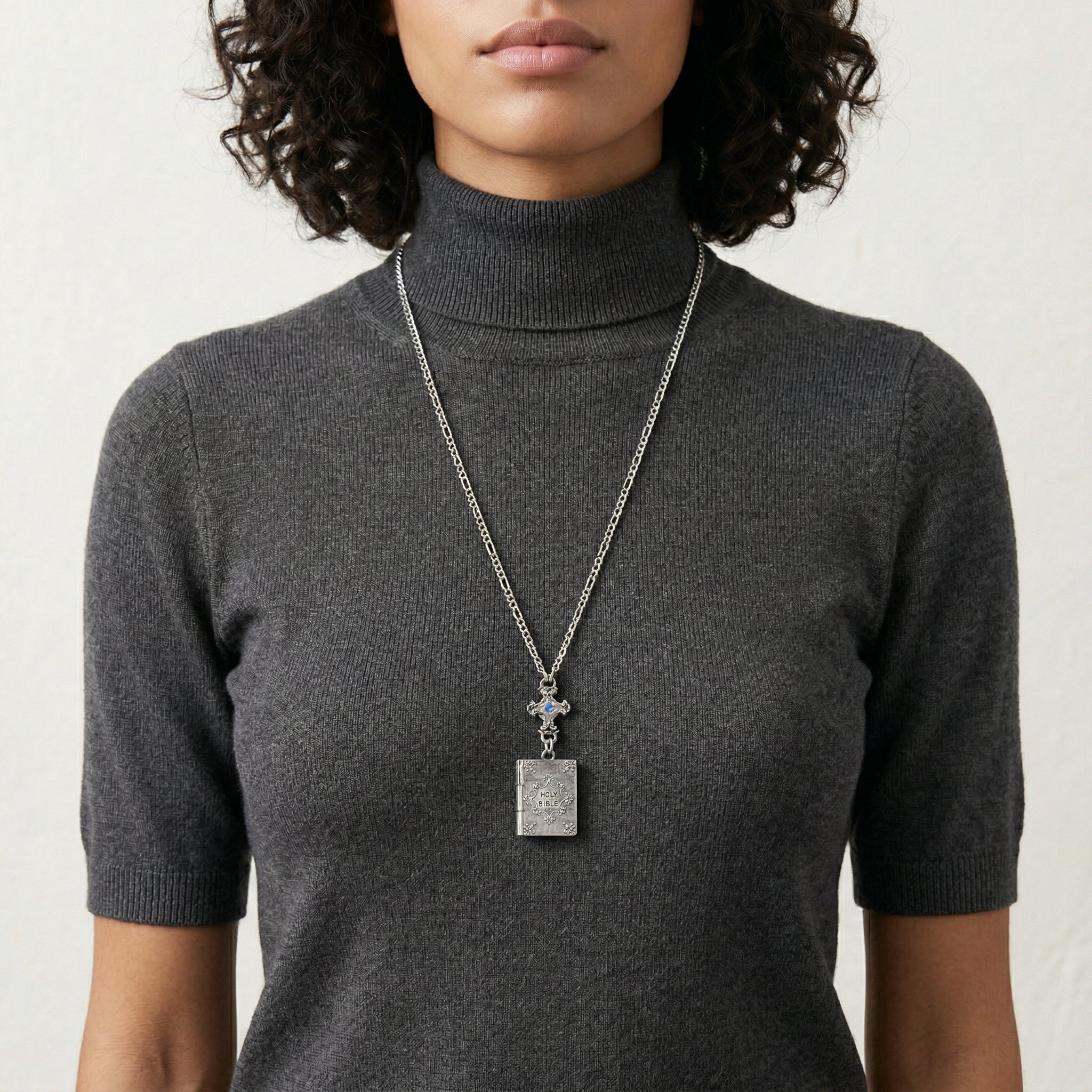Person wearing a dark gray turtleneck sweater with a long Holy Bible locket necklace.