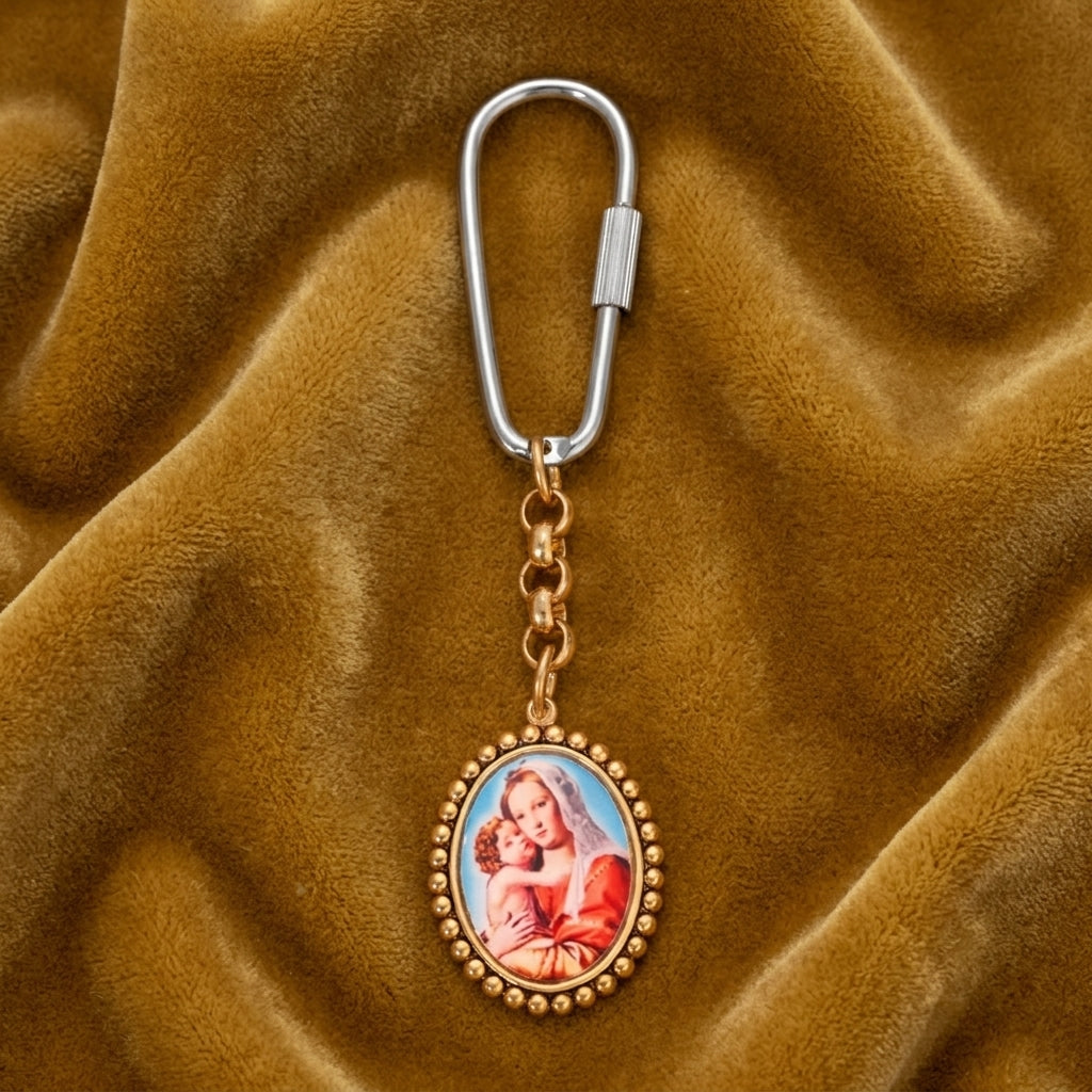 Oval gold tone Mother Mary and Child keychain with a carabiner resting on a dark yellow fabric surface