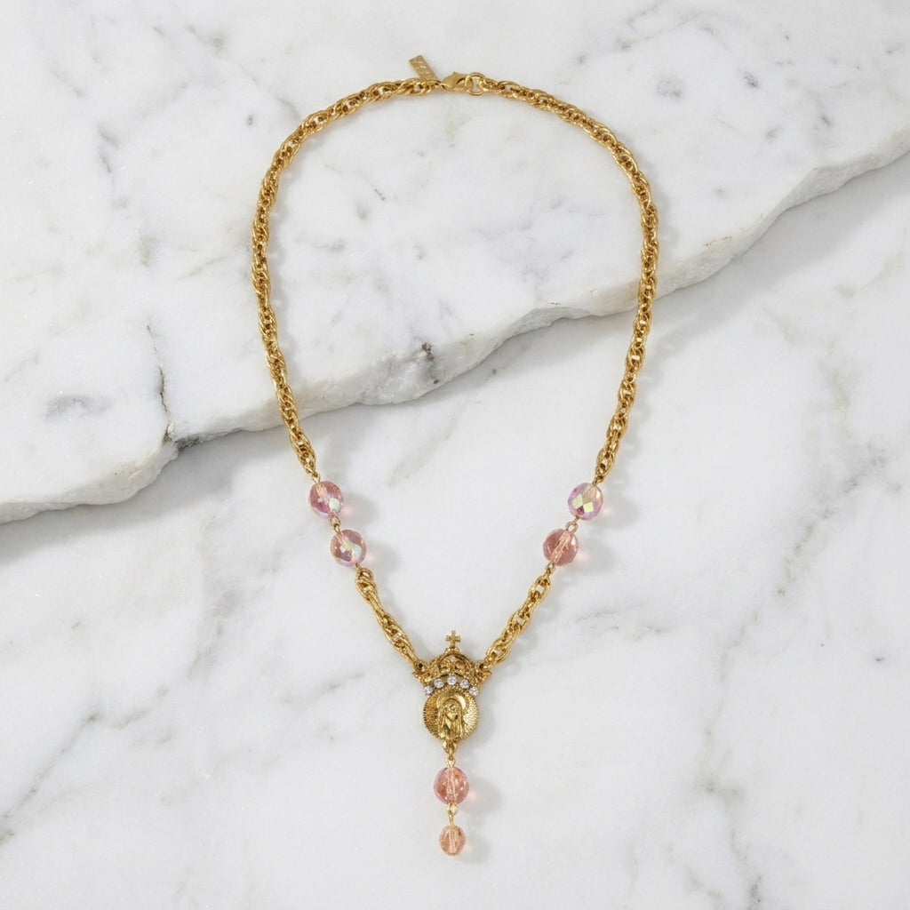Gold necklace with pink beads that has a cross, crown and Mother Mary pendant accented with clear crystals on a marble surface