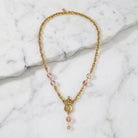 Gold necklace with pink beads that has a cross, crown and Mother Mary pendant accented with clear crystals on a marble surface