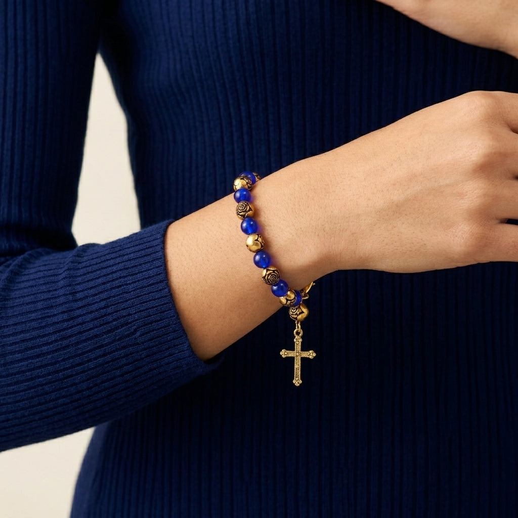 A close-up of a person's wrist wearing a Sapphire & Gold Rosebud Beaded Cross Charm Bracelet, paired with a dark navy ribbed sweater.