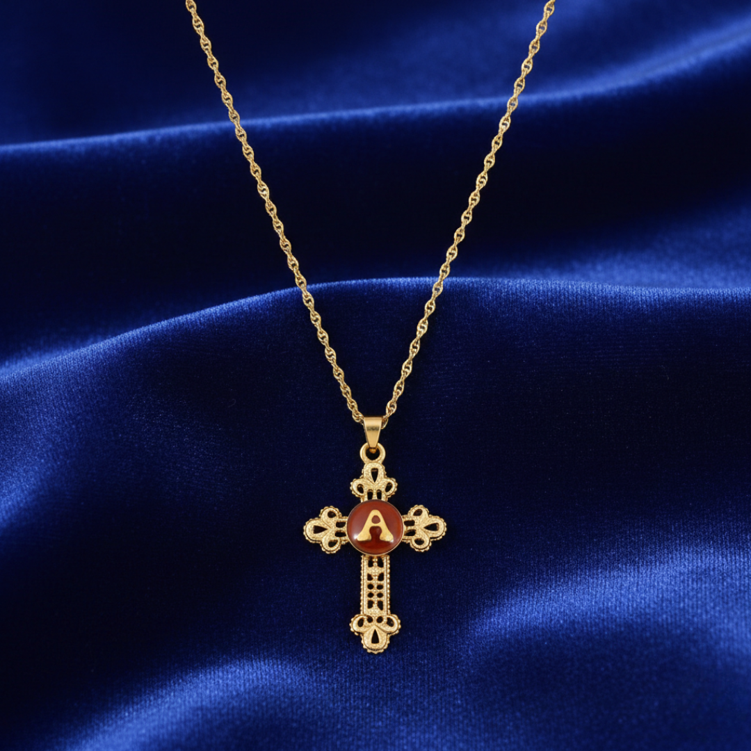Gold cross necklace initialed jewelry with a red stone on a blue satin background.