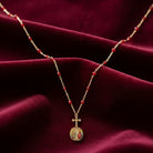 Gold necklace with red beads and a cross pendant featuring religious motifs on a burgundy fabric background