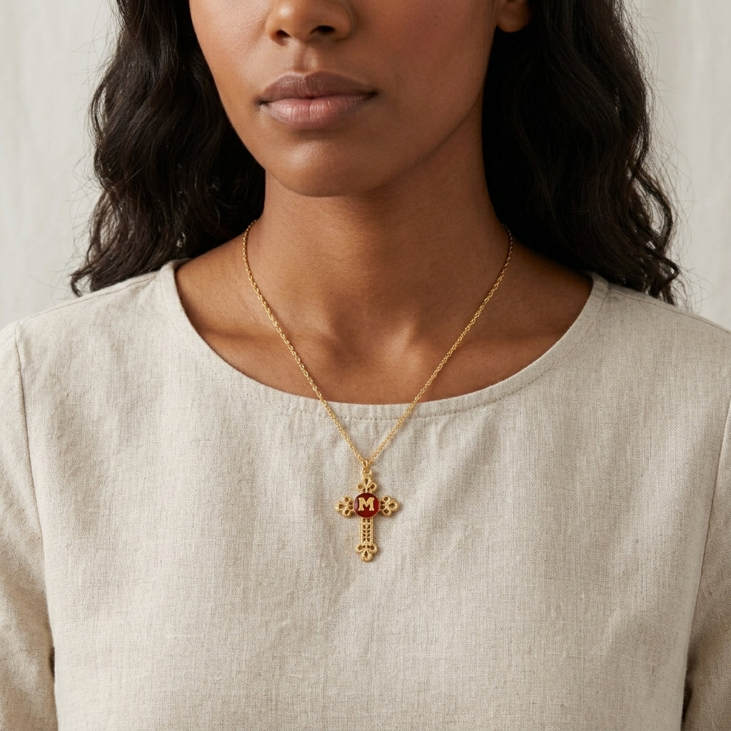 Woman wearing a gold necklace with am 'M' initialed cross pendant against a neutral background