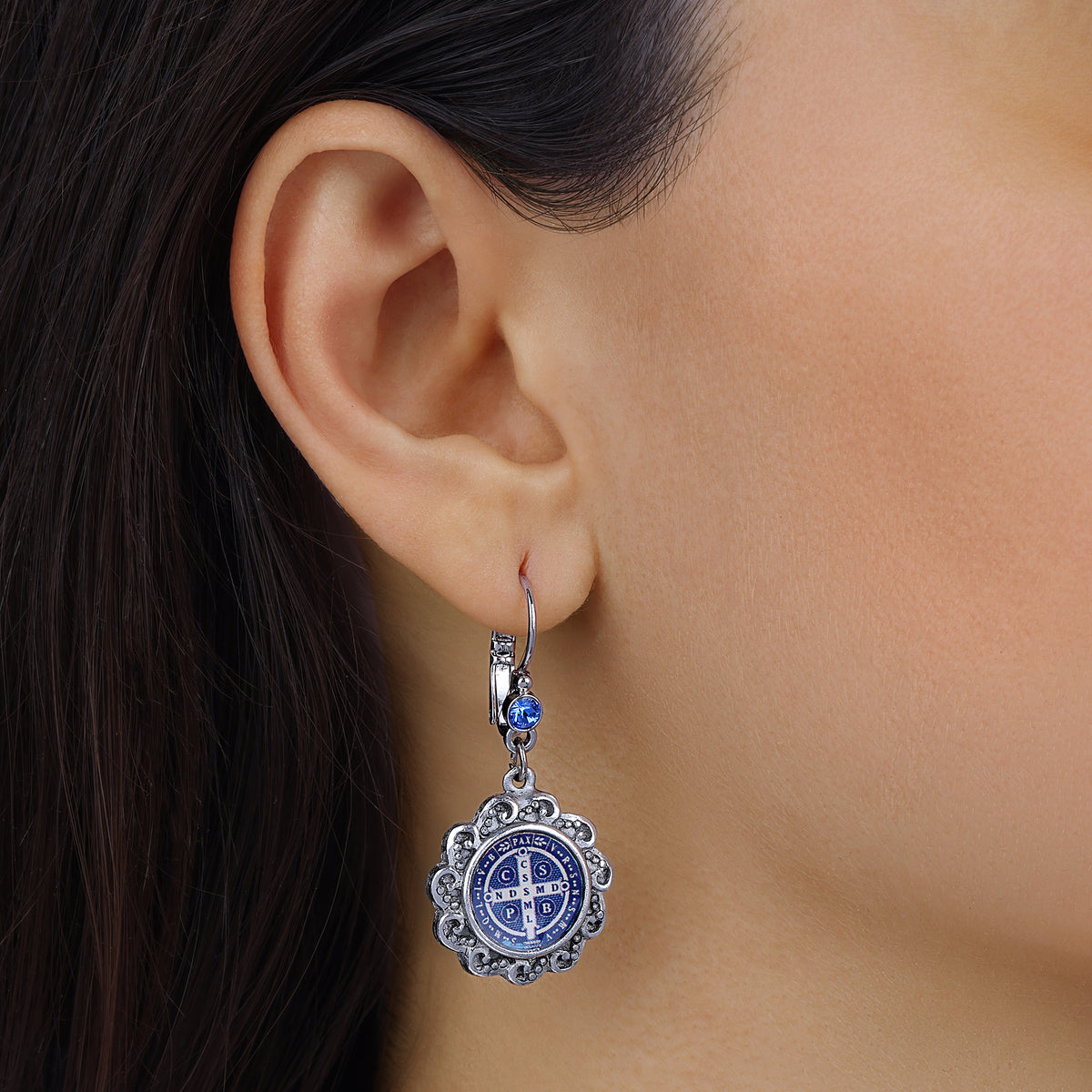 A close-up of a person's ear wearing a Round Silver & Blue Crystal Saint Benedict Decal Drop Earrings