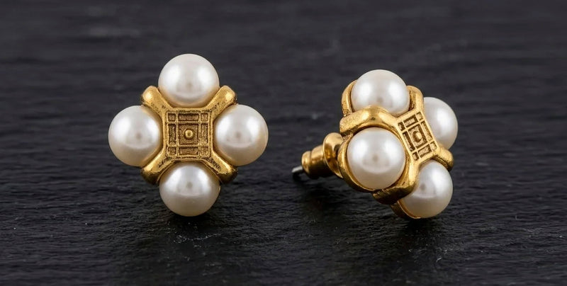 Vintage-style gold and pearl stud earrings on a dark textured slate background. Each earring features a central gold cross-shaped setting with a square medallion and four white pearl-like beads radiating from the center.